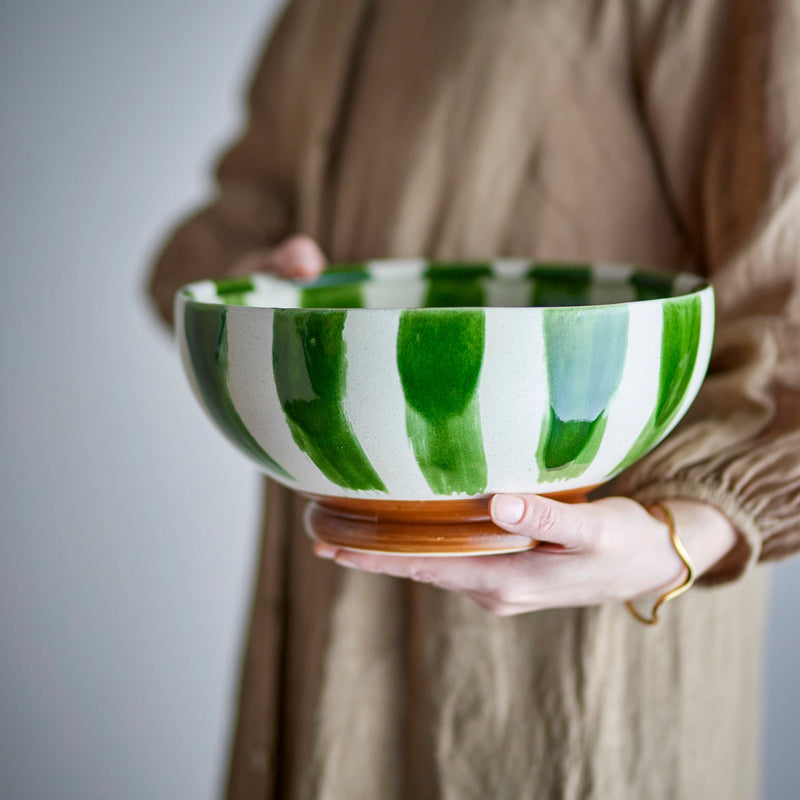 A person in a beige linen shirt holds a large ceramic bowl with vertical green and white stripes and a brown wooden base against a neutral gray background.