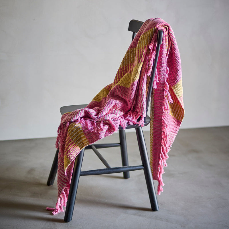 A pink and Lemon yellow striped knit throw blanket with fringed edges draped over a black wooden chair in a minimalist studio setting.
