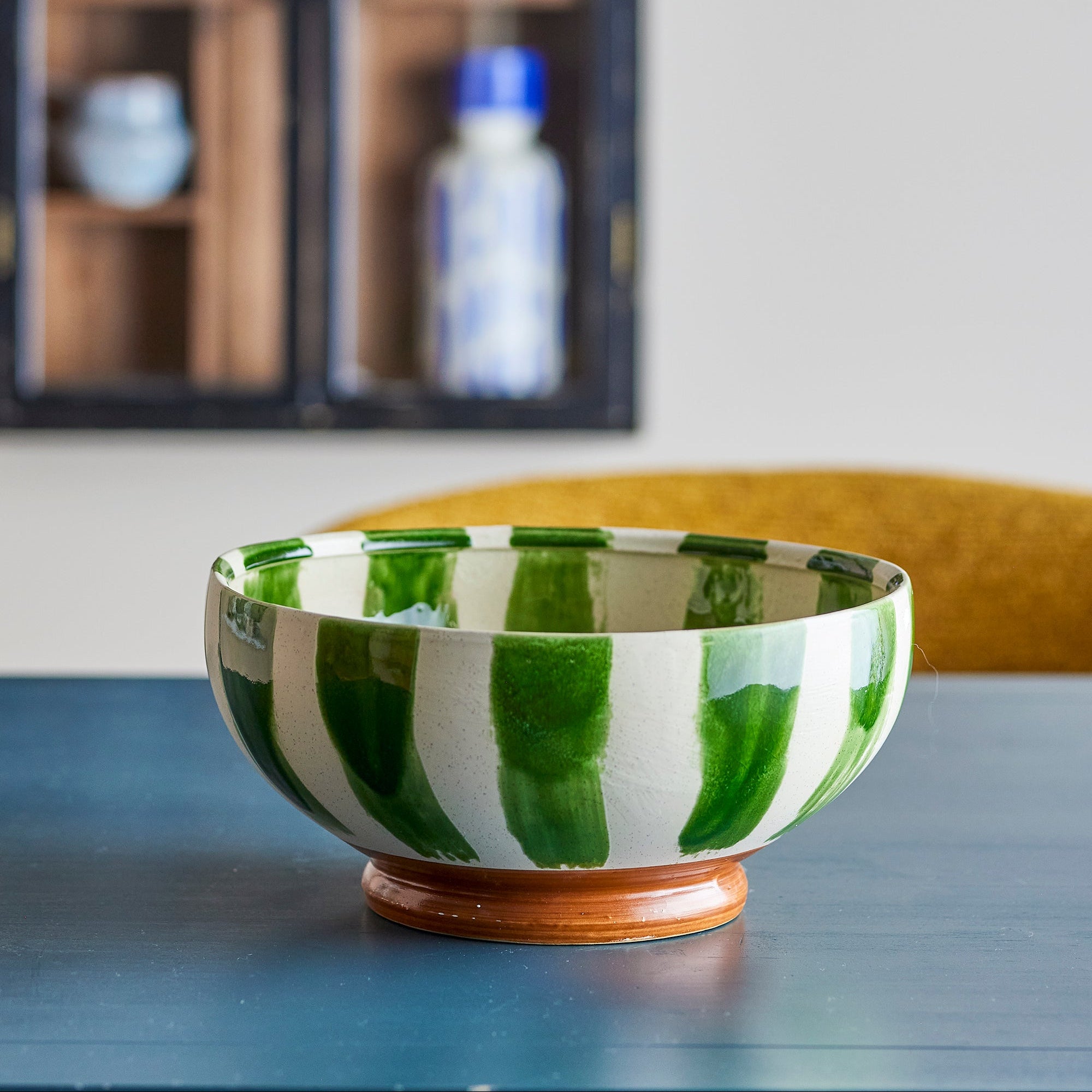 A ceramic bowl with green and white vertical brushstroke patterns and a brown wooden base, displayed on a dark table with a blurred background showing a shelf and chair.