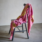 A pink and Lemon yellow striped knit throw blanket with fringed edges draped over a black wooden chair in a minimalist studio setting.