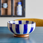 A ceramic bowl with bold vertical blue and white stripes and a small round wooden base, displayed on a dark tabletop with a blurred indoor background.