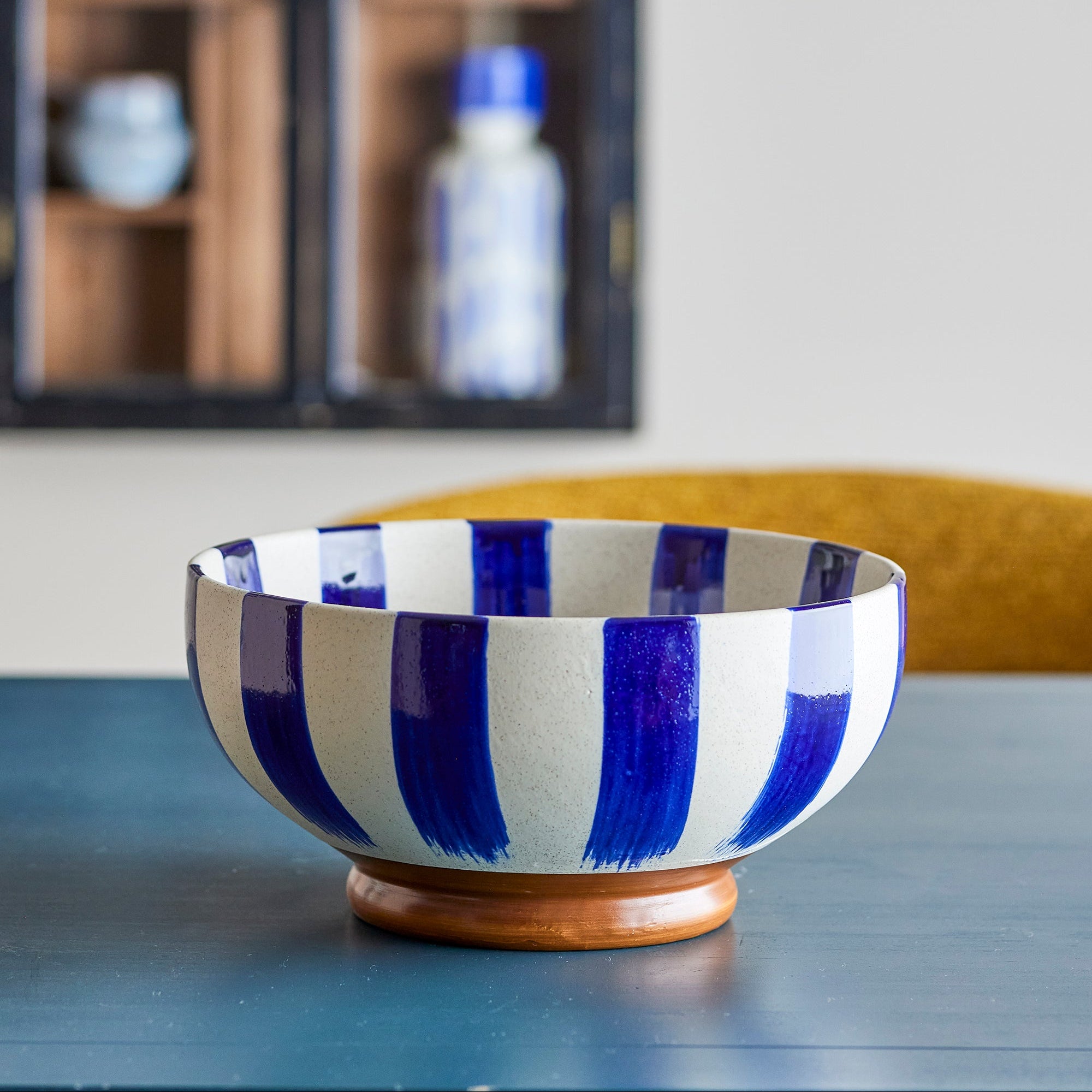 A ceramic bowl with bold vertical blue and white stripes and a small round wooden base, displayed on a dark tabletop with a blurred indoor background.