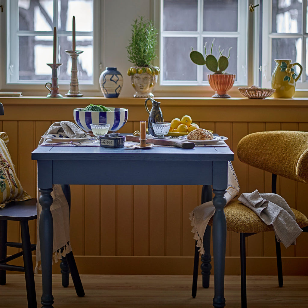 A styled dining room scene with a blue wooden table set with a striped bowl, plates, and candles, surrounded by yellow upholstered chairs in front of a window with a decorative windowsill.