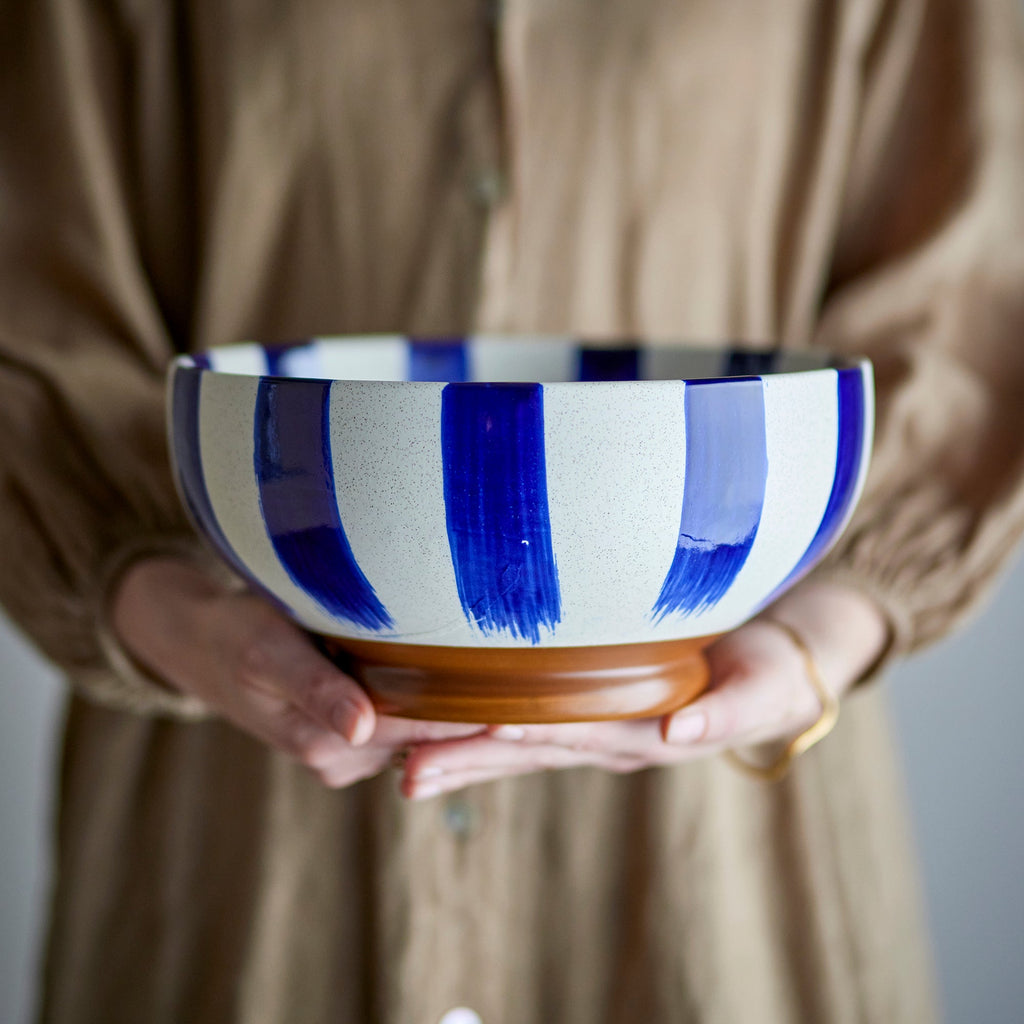 A person in a beige button-up shirt holds a ceramic bowl with wide vertical blue brushstroke stripes on a white body and a brown wooden base.