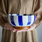 A person in a beige button-up shirt holds a ceramic bowl with wide vertical blue brushstroke stripes on a white body and a brown wooden base.