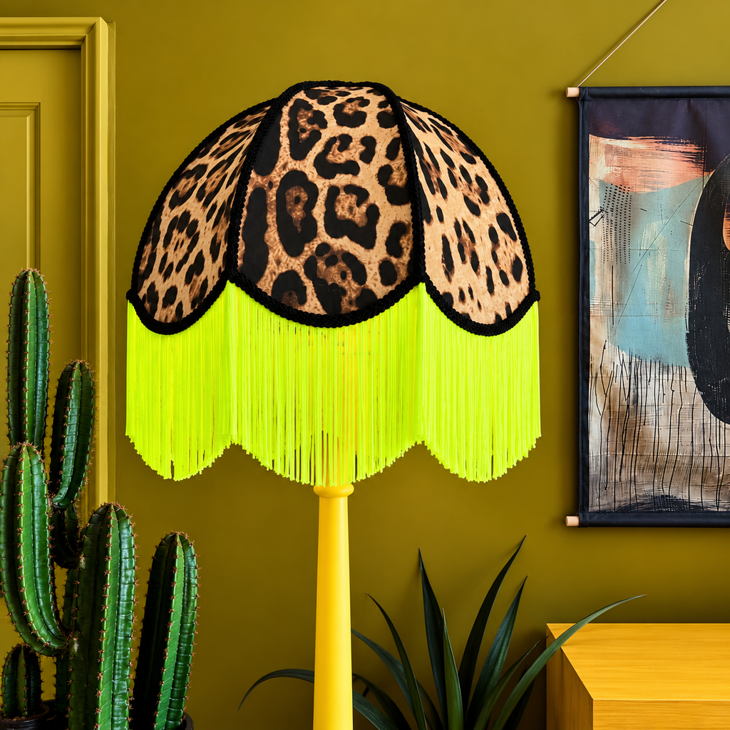 A decorative floor lamp with a leopard-print fabric shade and bright neon yellow fringe, set against a mustard yellow wall with a cactus and abstract wall art in the background.