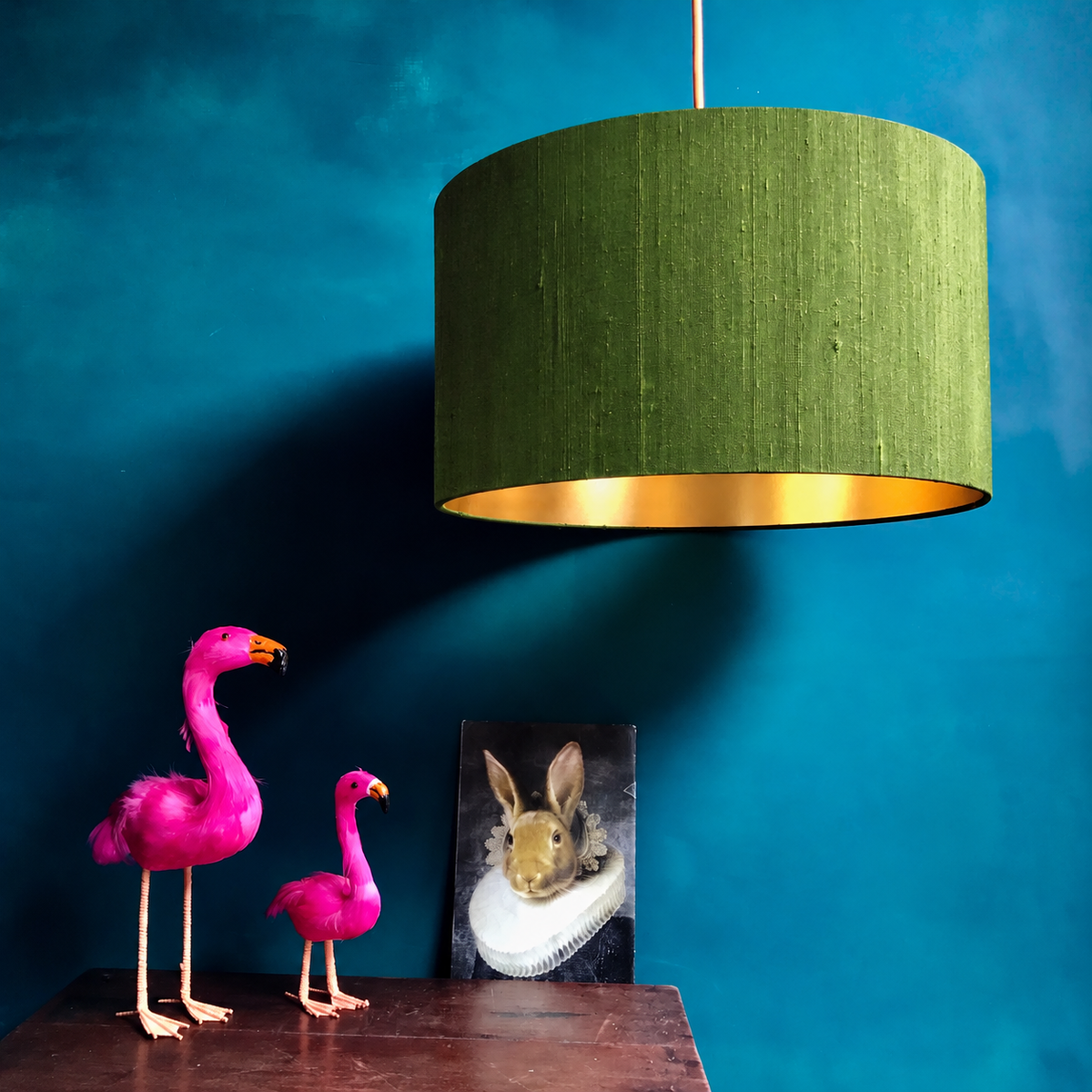 An olive green fabric drum pendant lamp with a gold interior lining hangs against a textured teal wall above a dark wooden table. On the table are two bright pink flamingo figurines and a small framed picture of a rabbit.
