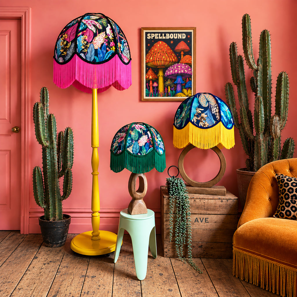 Three decorative table and floor lamps with colorful floral and butterfly-patterned shades and bright fringe, styled in a retro room with a pink wall, cacti, a mustard chair, and a wooden crate.
