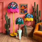 Three decorative table and floor lamps with colorful floral and butterfly-patterned shades and bright fringe, styled in a retro room with a pink wall, cacti, a mustard chair, and a wooden crate.