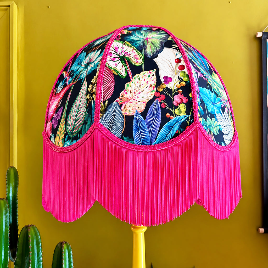 A dome-shaped lampshade with a vibrant tropical leaf and floral print on a black background, finished with a long bright pink fringe and mounted on a yellow pole against a mustard yellow wall.
