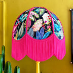 A dome-shaped lampshade with a vibrant tropical leaf and floral print on a black background, finished with a long bright pink fringe and mounted on a yellow pole against a mustard yellow wall.