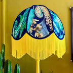 A decorative table lamp with a dome-shaped shade featuring a tropical leaf and floral print in blue, teal, and cream, with a long bright yellow fringe around the bottom edge. The lamp is shown against a mustard yellow wall with green cactus plants at the side.
