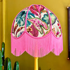 A decorative dome-shaped lampshade with a tropical leaf and fruit print on a cream background, finished with bright pink fringe and trim, mounted on a yellow lamp post against a mustard yellow wall.