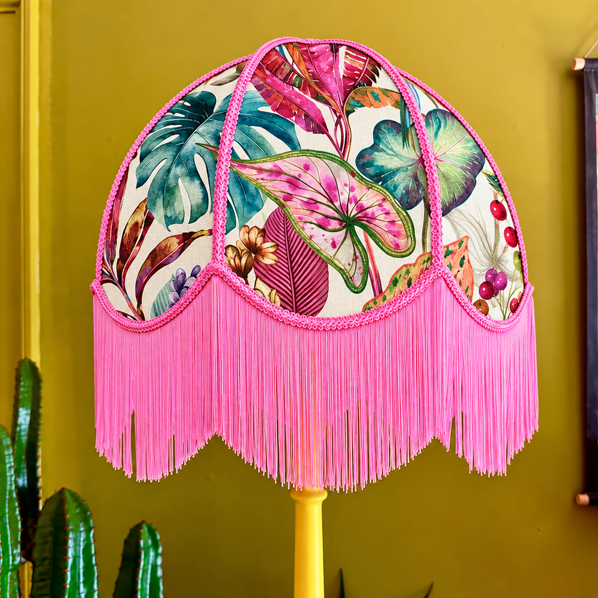 A decorative dome-shaped lampshade with a tropical leaf and fruit print on a cream background, finished with bright pink fringe and trim, mounted on a yellow lamp post against a mustard yellow wall.