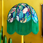 A decorative dome lampshade with a tropical leaf print on a navy background and a long green fringe hem, mounted on a yellow lamp against a mustard yellow wall.